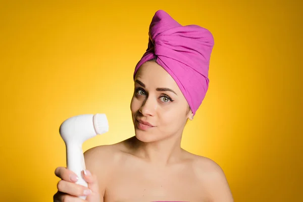 Ένα χαριτωμένο νεαρή κοπέλα με μια ροζ πετσέτα στο κεφάλι της κατέχει ένα ηλεκτρικό Βουρτσάκι για βαθύ καθαρισμό του προσώπου της — Φωτογραφία Αρχείου