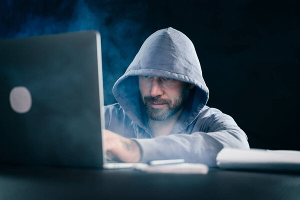 таинственный бородатый человек прячет лицо под капотом, взламывает ноутбук, в темноте
