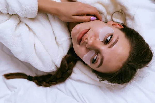 Una linda joven con una túnica blanca yace en una cama, bajo los ojos de un parche —  Fotos de Stock