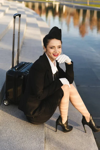 행복 한 젊은 여자 스튜 어디 스 제복에서 항공편을 기다리는 단계, 가방에 앉아 있다 — 스톡 사진