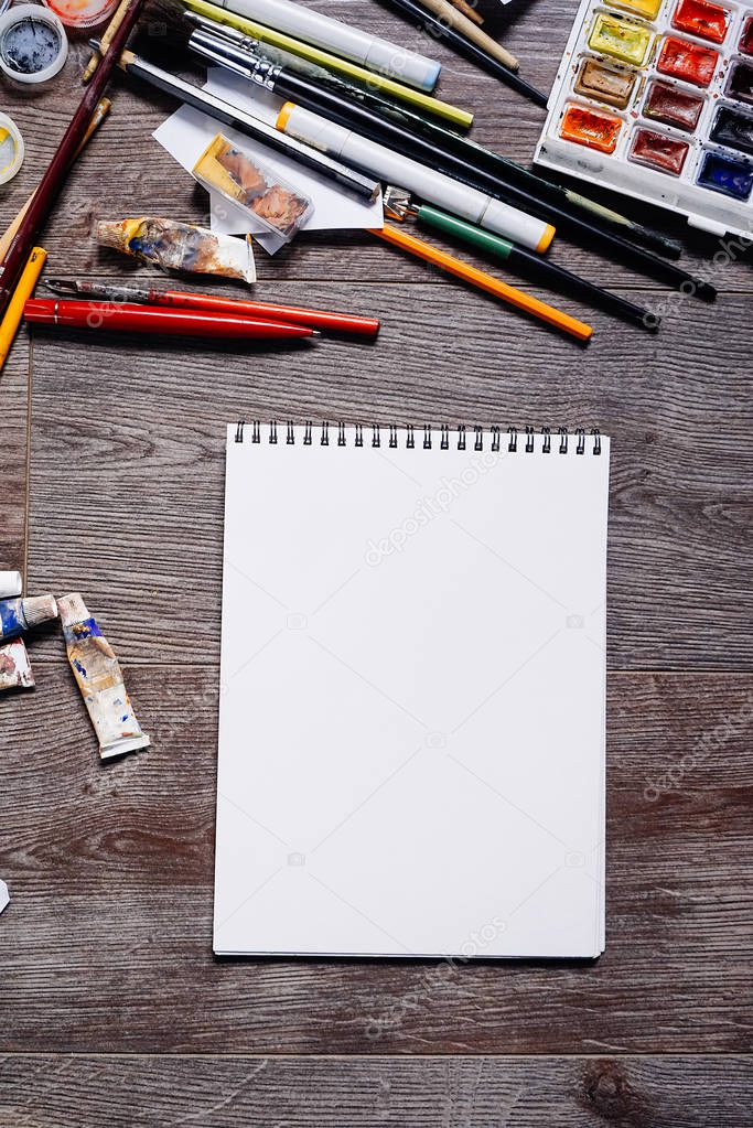 on a wooden surface is a white notepad for drawing, artistic paints and brushes