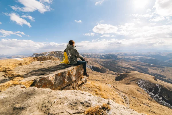 Jovem viajante senta-se na beira do penhasco, goza da natureza da montanha e do sol — Fotografia de Stock