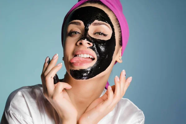 Αστείο κορίτσι με μια ροζ πετσέτα στο κεφάλι της και δείχνει μια γλώσσα, στο πρόσωπό της ένα μαύρο καθαρισμού μάσκα — Φωτογραφία Αρχείου