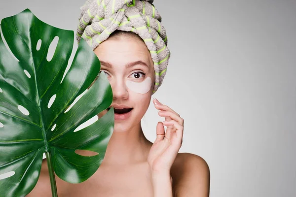 Όμορφη κοπέλα έκπληξη με μια πετσέτα στο κεφάλι της, κάτω από τα μάτια μπαλώματα, κρατά ένα πράσινο φύλλο, ημερήσιο σπα — Φωτογραφία Αρχείου