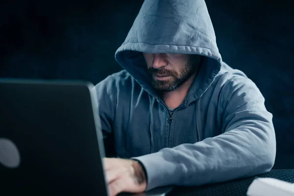 Criminoso barbudo homem esconde seu rosto sob o capô, hacks a senha no laptop — Fotografia de Stock