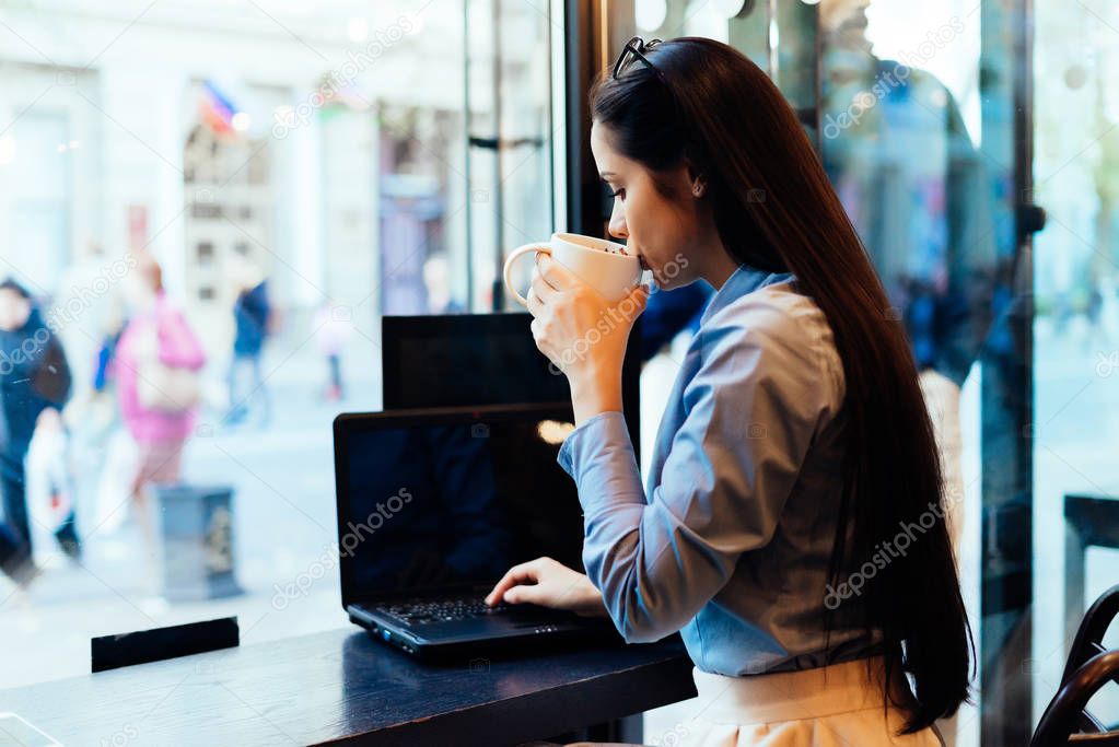 girl in a shirt with glasses on her head sits behind a laptop and drinks coffee