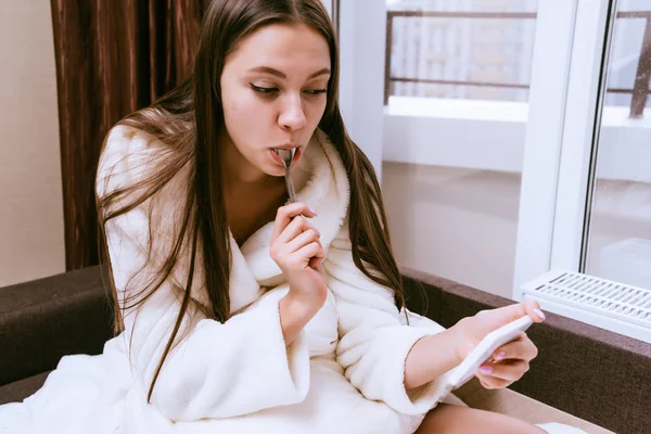 Menina de casaco branco senta-se na cama e olha para o telefone — Fotografia de Stock