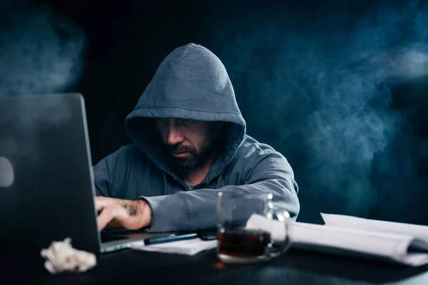 Ένας χάκερ σε ένα γκρι φούτερ κάθεται πίσω από ένα φορητό υπολογιστή — Φωτογραφία Αρχείου