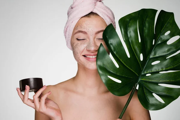 Feliz chica sonriente con una toalla rosa en la cabeza se aplica un exfoliante de café en la cara, spa de día — Foto de Stock