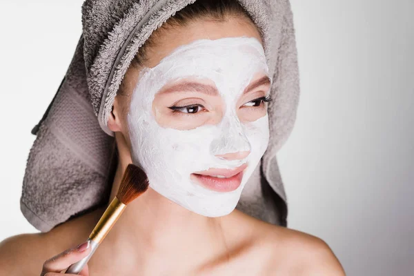 Jovem sorrindo menina com uma toalha na cabeça faz um tratamento de spa, coloca em um rosto uma máscara branca hidratante com uma escova — Fotografia de Stock
