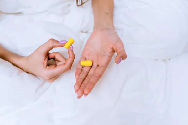 Dziewczyna siedzi na łóżku pod biały koc, trzymając żółty zatyczki do uszu przed hałasem — Zdjęcie stockowe