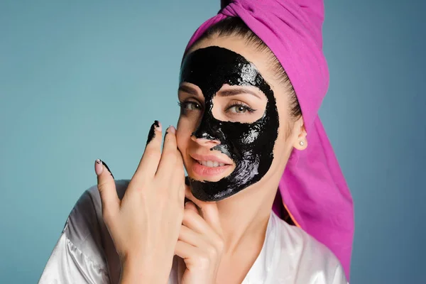 Menina sorridente bonito com uma toalha rosa na cabeça aplicar uma máscara preta contra a acne no rosto — Fotografia de Stock