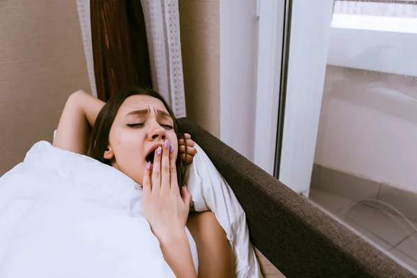 Сонная девушка лежит в постели под белым одеялом, зевает — стоковое фото