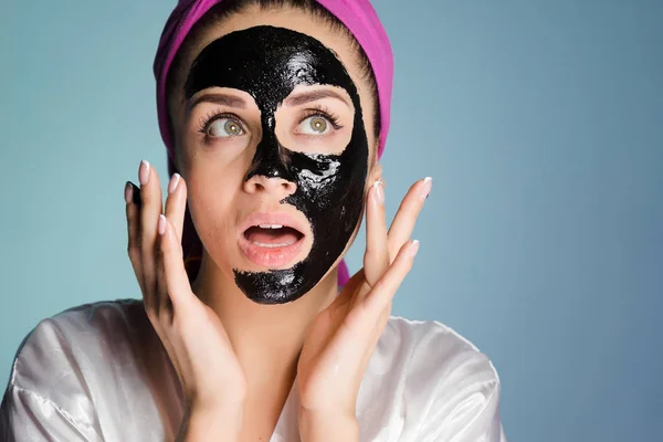 Joven sorprendida chica con una toalla rosa se aplica una máscara de limpieza negro en la cara — Foto de Stock