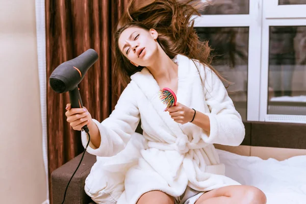Girl in white bathrobe dries hair with a hairdryer after shower — Stockfoto
