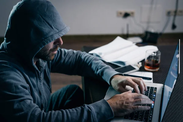 Guy hacker in a sweatshirt with a hood prints on a laptop — Stock Photo, Image