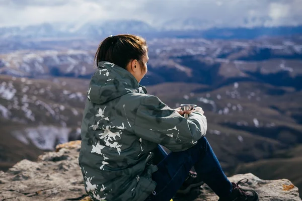 Chica turista en una chaqueta se sienta en el fondo de las montañas y bebe té —  Fotos de Stock