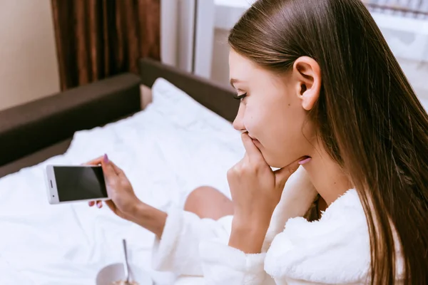 Gadis muda duduk di tempat tidur menonton video di telepon — Stok Foto