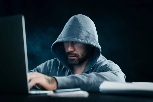 Jovem hacker em uma camisola com um capuz senta-se atrás de um laptop — Fotografia de Stock