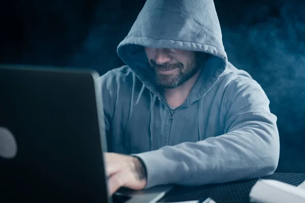 Cara hacker em uma camisola com um capuz senta-se atrás de um laptop — Fotografia de Stock