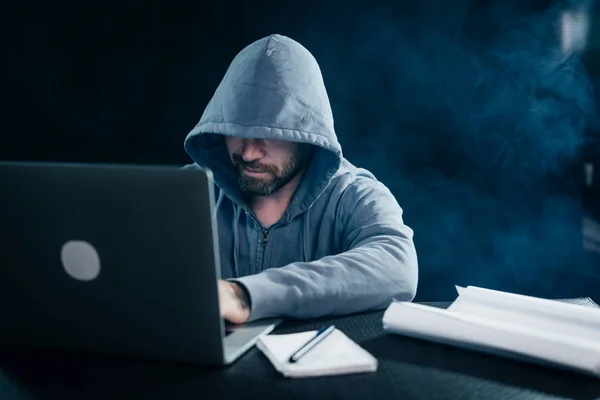 Um jovem hacker trabalhando atrás de um laptop em um capuz — Fotografia de Stock