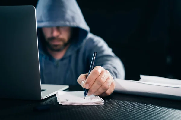young hacker making notes in a notebook sitting behind laptop