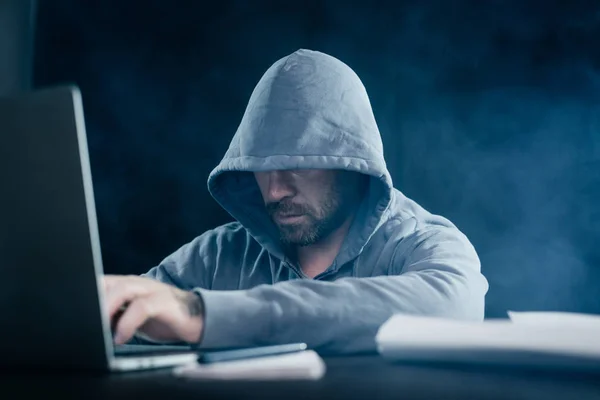 Um hacker experiente trabalha atrás de um laptop em um capuz — Fotografia de Stock