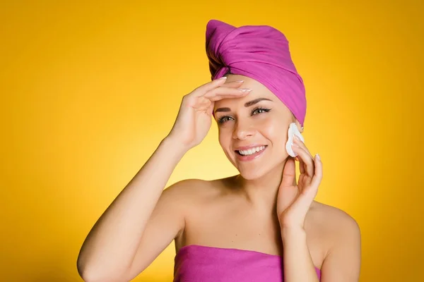 Ευτυχισμένος χαμογελαστό κορίτσι με μια ροζ πετσέτα στο κεφάλι της καθαρίζει το δέρμα στο πρόσωπό της με wadded λευκό δίσκο — Φωτογραφία Αρχείου
