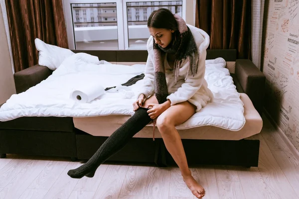 Sleepy young girl in a bathrobe sits on the bed and puts on warm socks — Stock Photo, Image