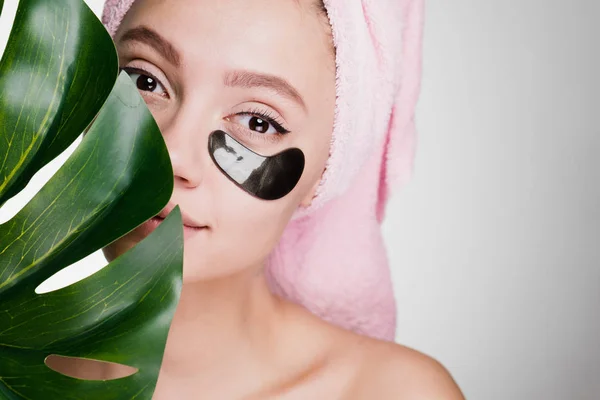 可爱的年轻女孩用粉红色的毛巾在她的头上持有绿叶, 在黑色补丁的眼睛, 日间水疗 — 图库照片