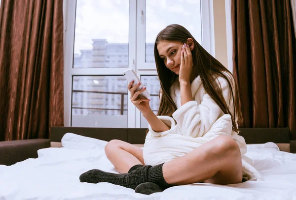 Χαριτωμένο νεαρή κοπέλα σε ένα λευκό χιτώνα και ζεστές κάλτσες κάθεται στο κρεβάτι και κοιτάζει σε ένα smartphone — Φωτογραφία Αρχείου