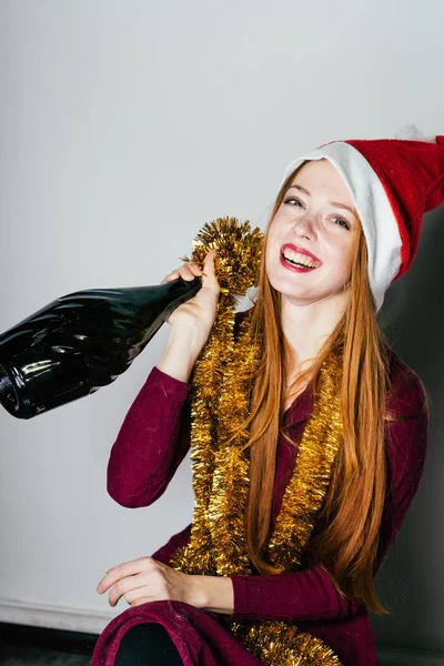 Redhead šťastná dívka v červené čepici jako Santa Claus slaví Nový rok drží láhev šampaňského — Stock fotografie