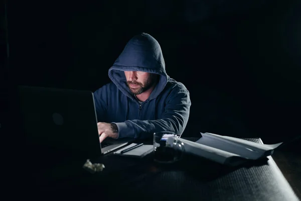 Εγκληματική γενειοφόρος άνδρας κρύβει το πρόσωπό του κάτω από την κουκούλα, κάθεται στο σκοτάδι, αμυχές του φορητού υπολογιστή — Φωτογραφία Αρχείου