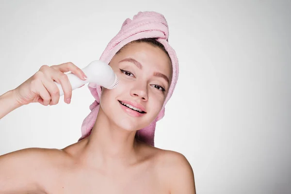 A cute smiling girl with a towel on her head doing a deep cleansing of the skin on her face using an electric brush — Stock Photo, Image