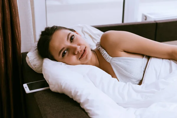 Милая девушка лежит в постели под белым одеялом, рано проснувшись — стоковое фото