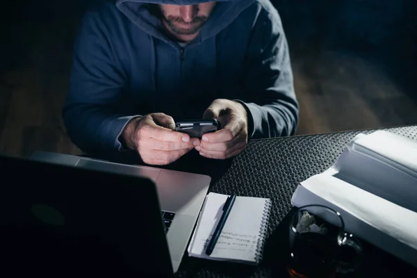 A criminal bearded man in a hood hacks a password on a smartphone, in the dark — Stock Photo, Image