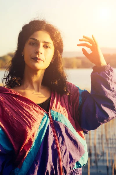 Girl with dark hair and a colorful jacket posing against the lake — Stock Photo, Image