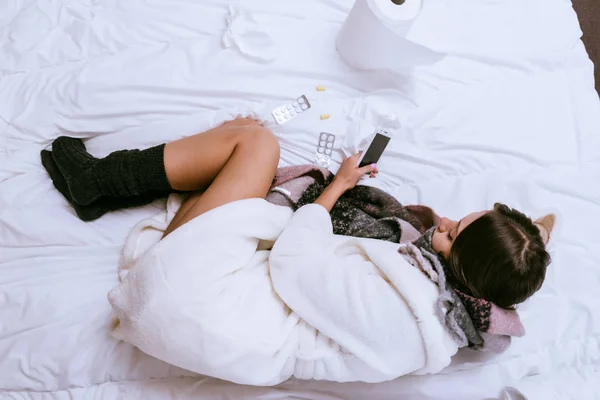 Una ragazza con un raffreddore in una vestaglia calda e calzini è sdraiata in un letto — Foto Stock