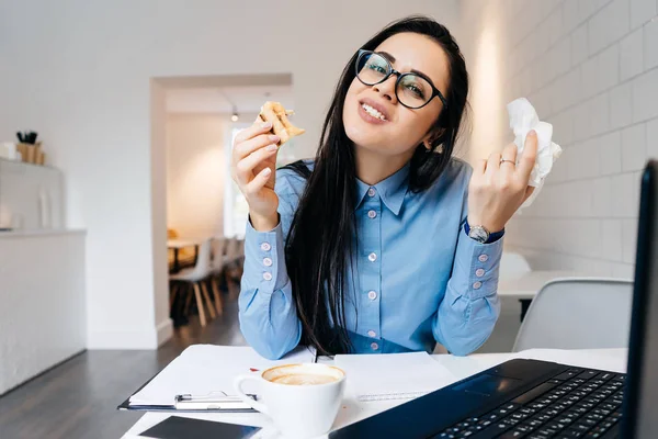Divertida joven con camisa azul y gafas almorzando en la cafetería después del trabajo —  Fotos de Stock