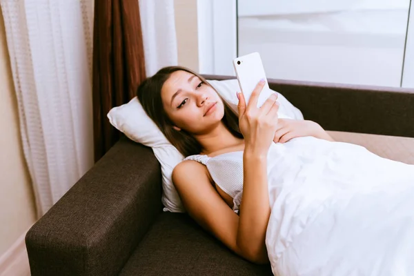 Милая сонная девушка лежит в постели, рано утром, смотрит в свой смартфон — стоковое фото