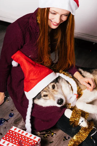 Laughing red-haired girl in a red cap sits on the floor with her dog, celebrates the new year — Stock Photo, Image