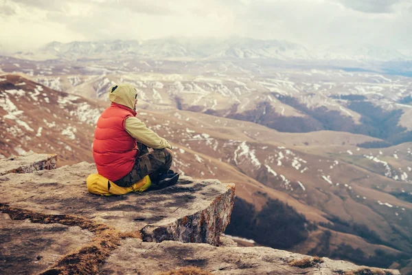 Viajero masculino en una chaqueta roja se sienta en el borde de un acantilado, en una mochila amarilla, disfruta de la naturaleza —  Fotos de Stock
