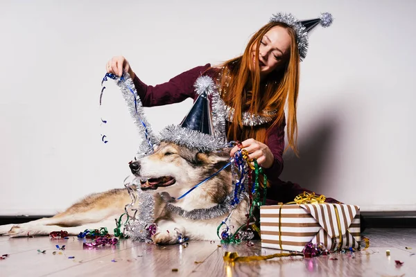 Mignonne fille aux cheveux roux avec une casquette sur la tête s'assoit sur le sol avec son chien, célèbre la nouvelle année et Noël, argent tinsel et cadeaux — Photo