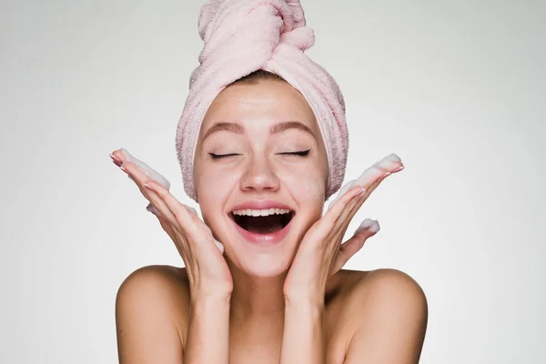 彼女の頭の上のピンクのタオルで素敵な笑っている女の子は彼女の顔に洗顔フォームを適用します。 — ストック写真