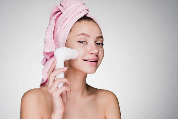 Mignonne fille souriante avec une serviette rose sur la tête nettoie la peau sur son visage avec une brosse électrique — Photo
