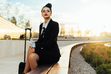 düşünceli genç kadın hostes üniformalı ve bir çanta ile parkta oturur ve onu uçuş için bekler