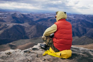 Kırmızı ceketli erkek bir gezgin üzerinde sarı bir sırt çantası bir uçurumun kenarına oturur ve temiz dağ havası sahiptir