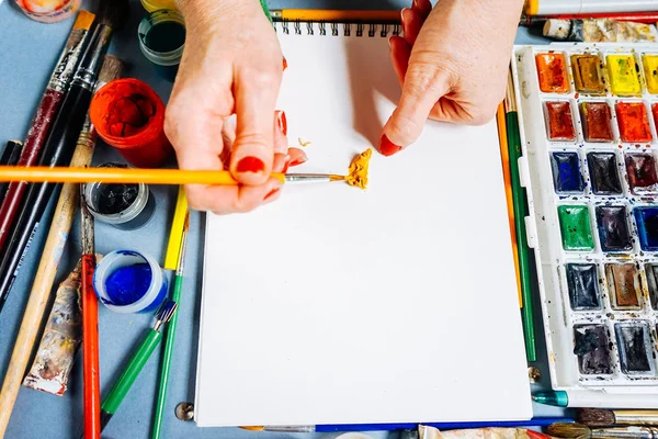 Μια γυναίκα να χρωματίζει ένα σχέδιο στη Λευκή Βίβλο με μια βούρτσα, δίπλα σε αυτό υπάρχει μια πολύχρωμα γκουάς και ακουαρέλες — Φωτογραφία Αρχείου