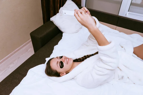 Menina feliz no manto branco jaz na cama, sob os olhos de manchas pretas, faz selfie — Fotografia de Stock