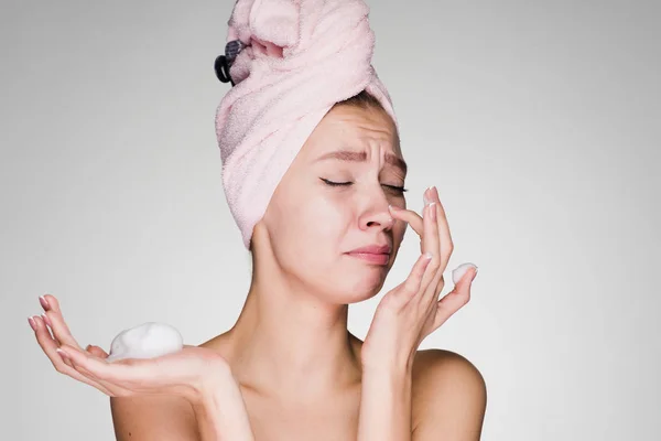 Funny young girl with a pink towel on her head applies a cleansing foam on her face — Stock Photo, Image